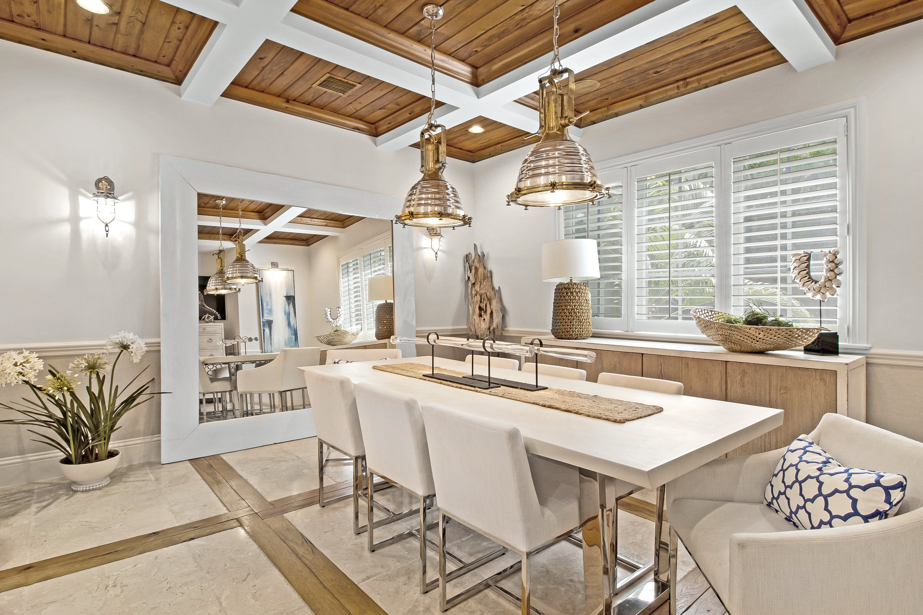 A bright, modern dining room with a white rectangular table, six white bar stools, a white dining chair in the foreground, a blue-and-white patterned pillow, wood ceiling beams, and large windows with white shutters.