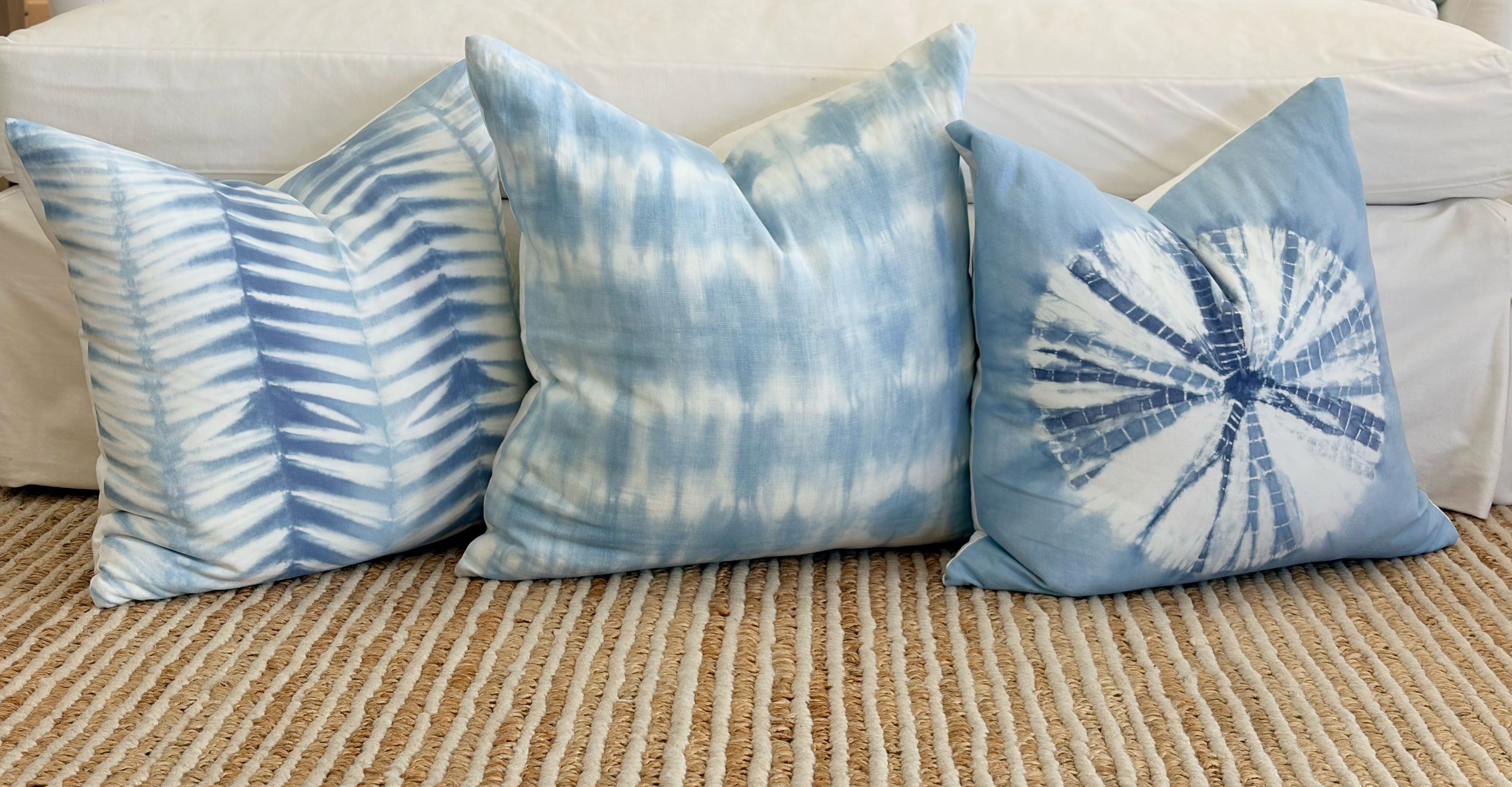 Four blue and white tie-dye throw pillows arranged on a woven beige rug in front of a white sofa.