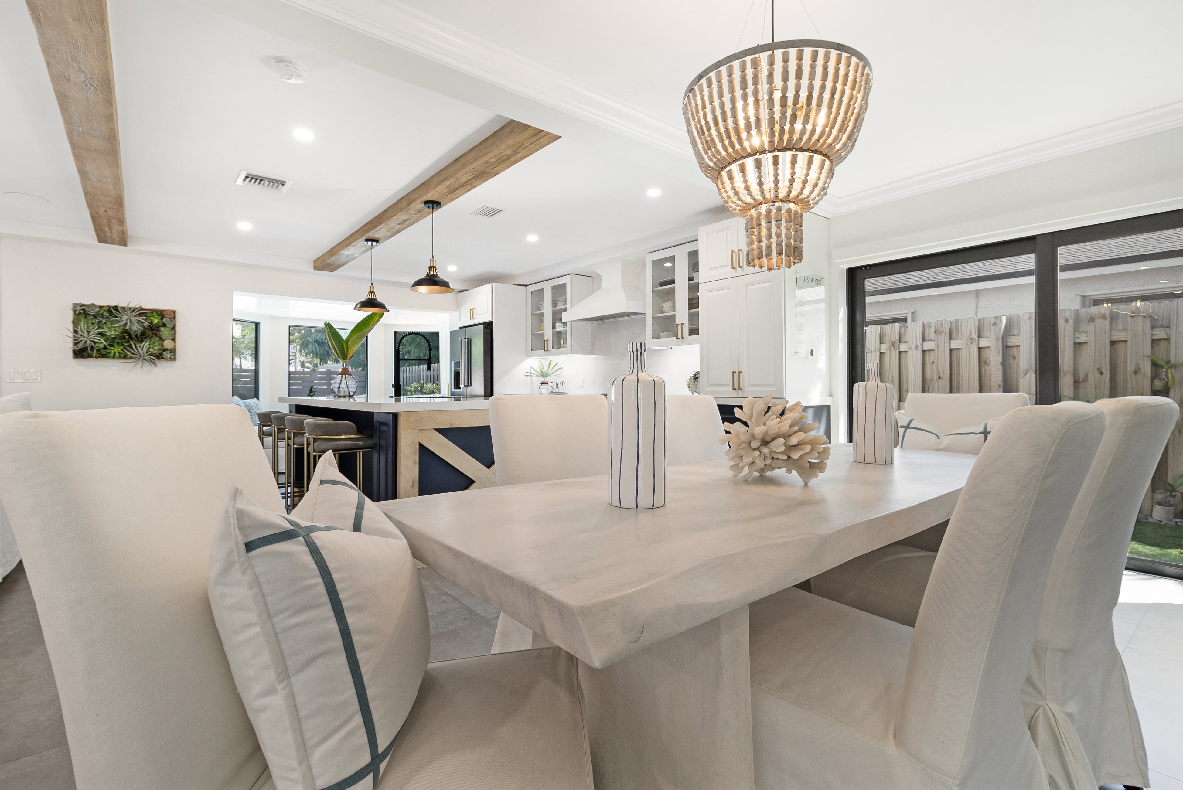 A bright, modern dining room featuring a light wood rectangular table surrounded by white upholstered dining chairs with blue stripe accents. A large woven natural-fiber chandelier hangs over the table, and a contemporary kitchen is visible in the background.